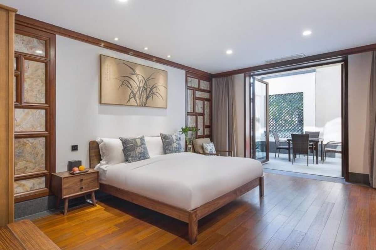 A modern bedroom with a large bed, wooden flooring, a nightstand, a wall-mounted artwork, and a sliding glass door leading to a balcony with outdoor seating.