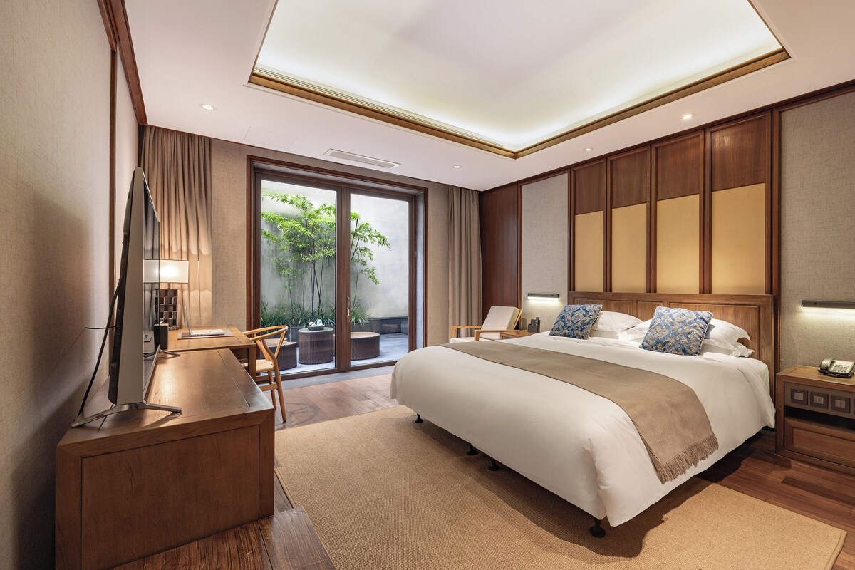 A modern hotel room with a large bed, a desk, a chair, a nightstand, and a view of greenery outside.