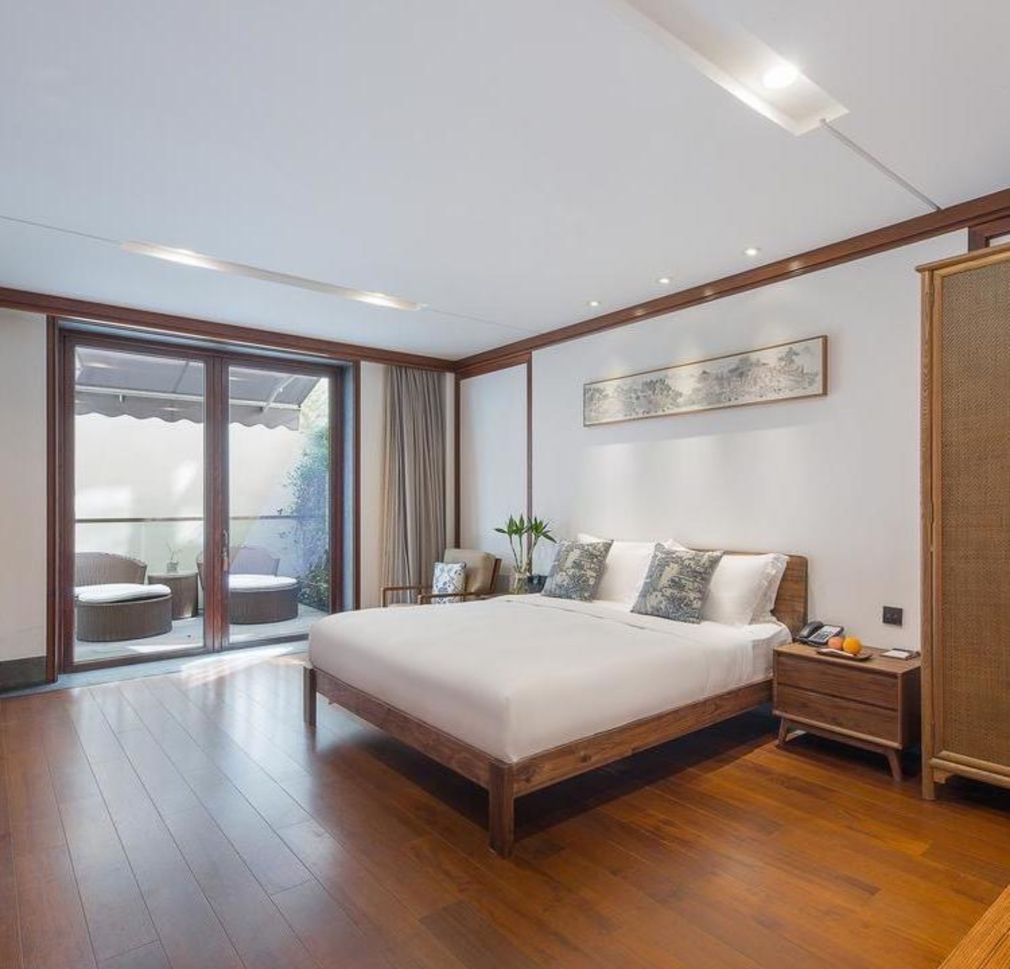 A modern bedroom with a large bed, wooden flooring, a sliding glass door to a balcony, and a small bedside table with a lamp.