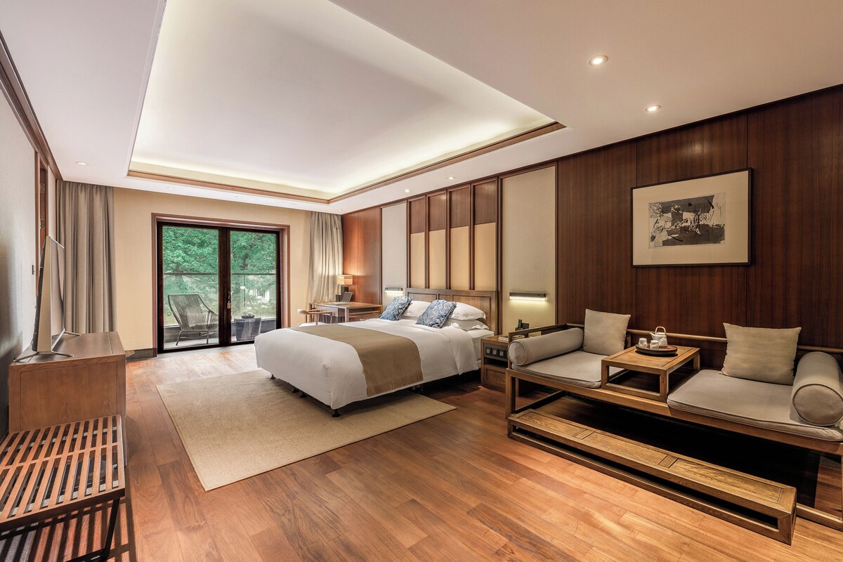 A modern hotel room with a large bed, a sofa, a desk, and a view of greenery through the sliding doors.