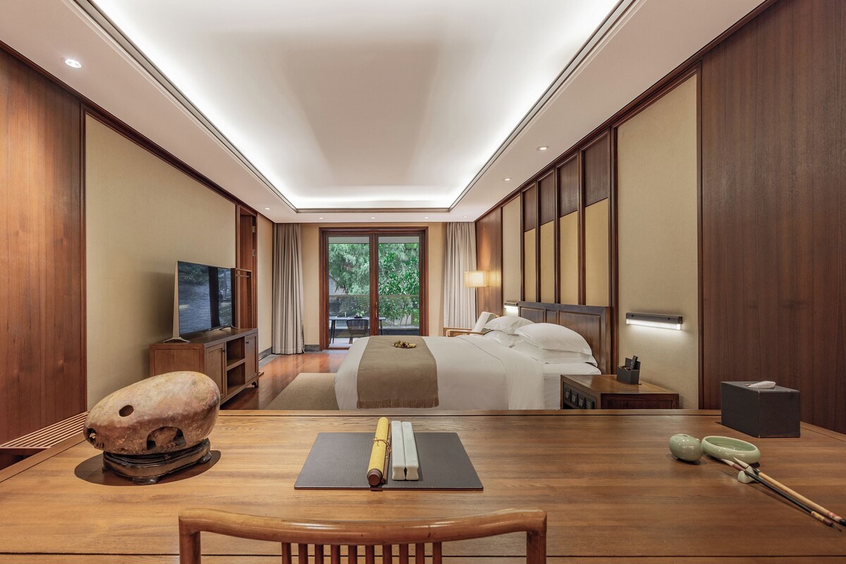 A hotel room with a bed, bedside table, TV, and a view of greenery outside.