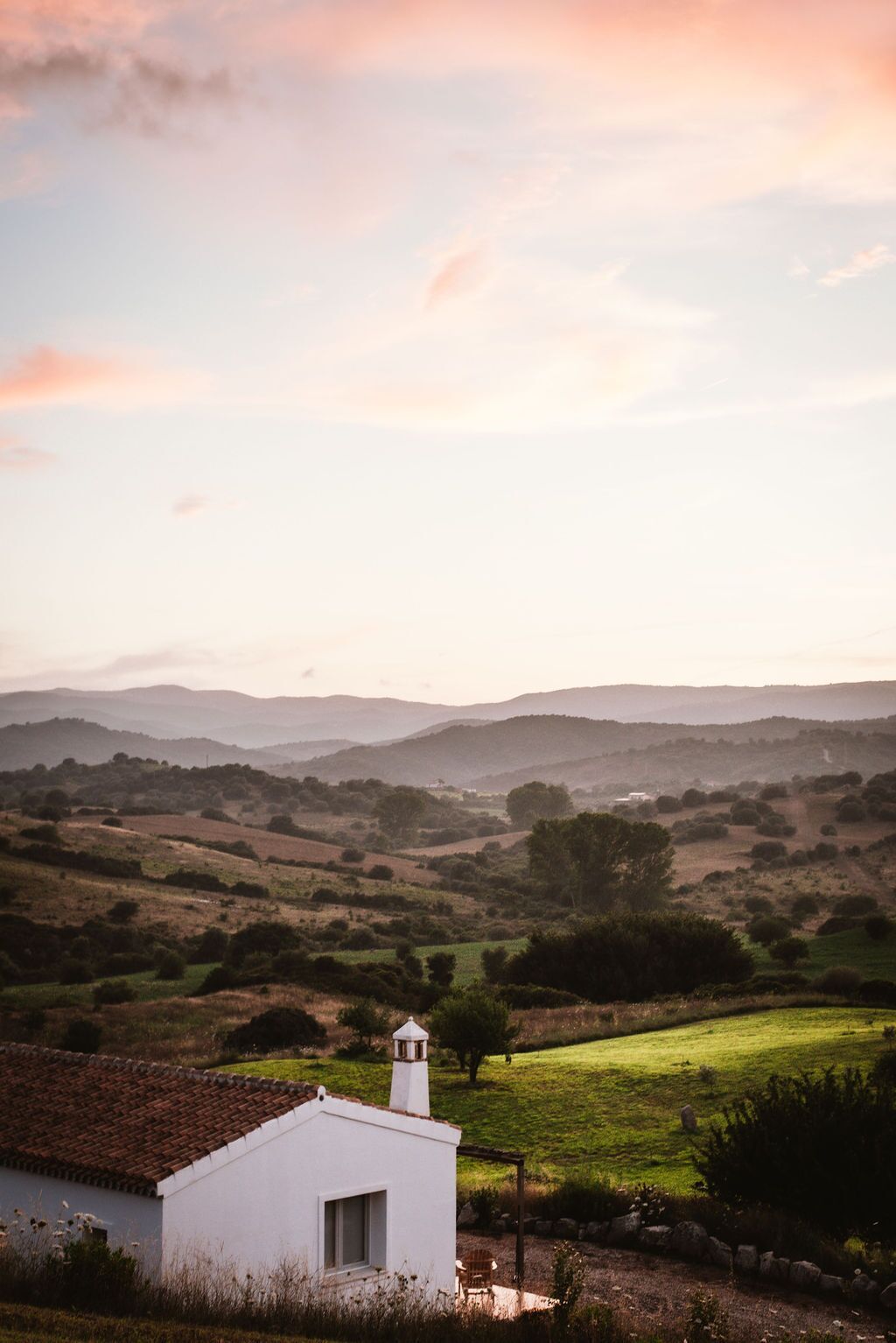 A white building with a tiled roof, surrounded by green fields and hills in the background.