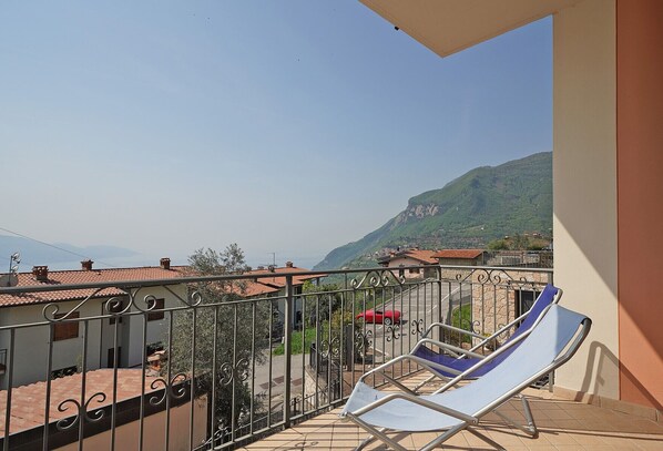 A balcony with a metal railing, a blue lounge chair, and a view of a mountainous landscape with buildings in the distance. - Luna: lake view apartment with two bedrooms (Tignale)