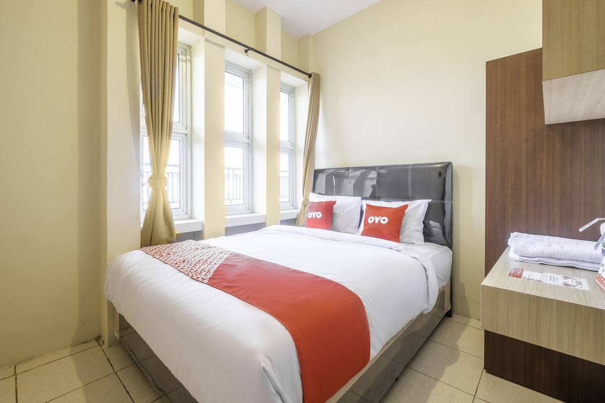 Ein Hotelzimmer mit Bett, Holz-Kopfteil, Fenster mit Vorhängen und einem kleinen Tisch mit Handtuch.