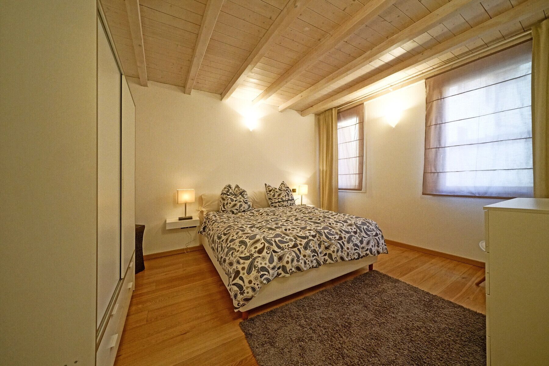 Un dormitorio con una cama, dos lámparas de noche, un ventanal con cortinas y techo de madera.