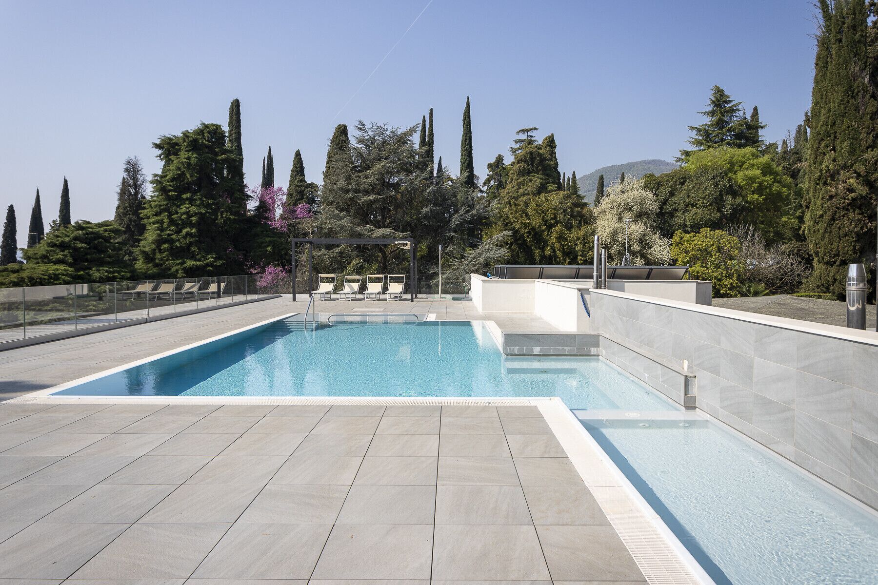 A rectangular swimming pool with a tiled deck, surrounded by trees and a clear sky.