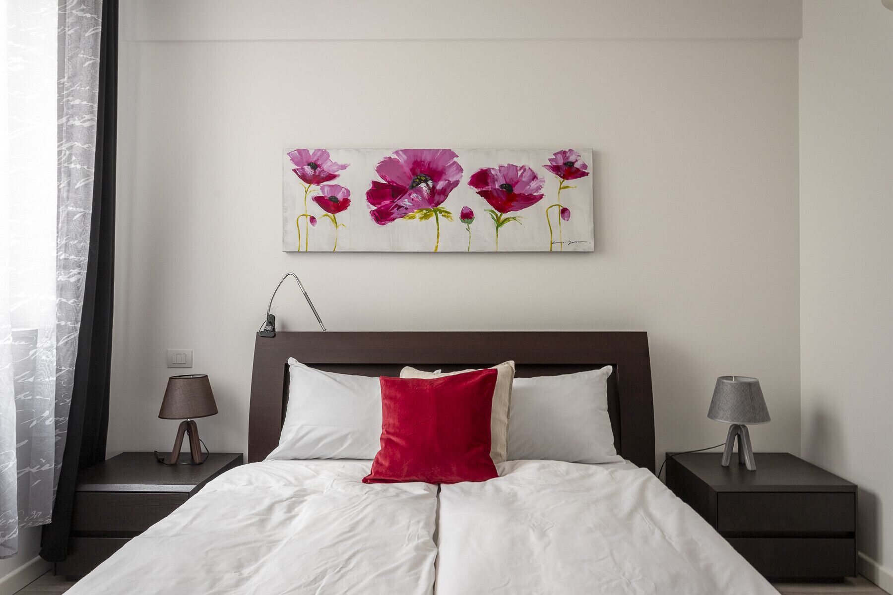 A bedroom with a bed, two bedside tables, a lamp on each table, a painting of flowers on the wall, and a window with sheer curtains.