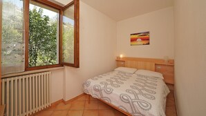 Un dormitorio pequeño con cama, radiador, ventana con vistas a zonas verdes y un cuadro en la pared.
