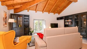 Een woonkamer met een hoog houten plafond, een bank, een gele fauteuil en een boekenkast.