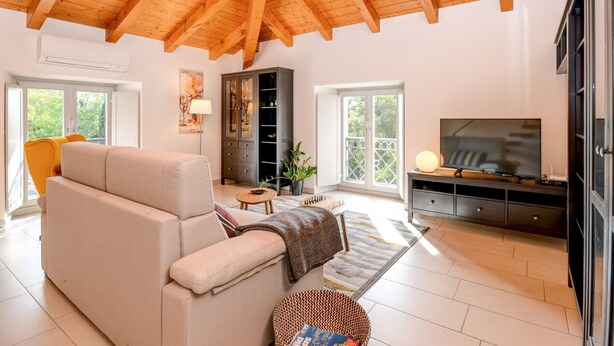 Een ruime woonkamer met een hoog houten plafond, een beige bank, een televisie en een kast.