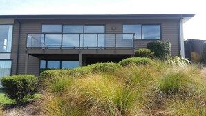 A modern two-story house with a balcony, surrounded by tall grasses and small shrubs.