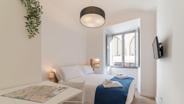 A minimalist hotel room with a bed, a desk with a map, a hanging plant, and a TV mounted on the wall. - Roma Station House (Rome)