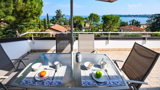 A glass table with a view of a coastal landscape, two chairs, and a table setting with plates, a bowl, a spoon, a fork, and a cup. - Cupido: with big lake view balcony in Gardone Riviera (Gardone Riviera)