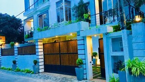 Un edificio moderno de dos pisos con entrada protegida, plantas en macetas y un balcón con vegetación.