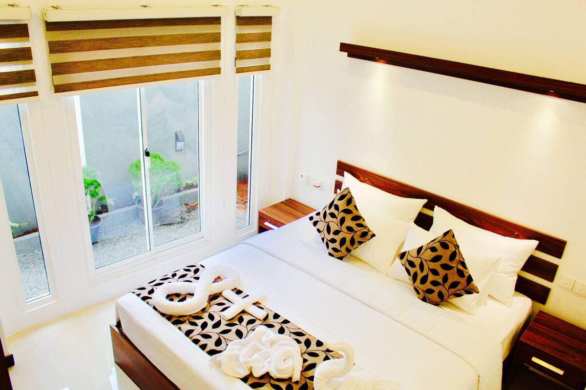 A hotel room with a large bed, multiple pillows, a bedside table, and a window with blinds.