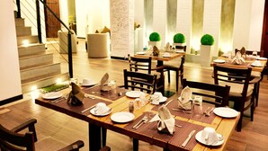 Un comedor con mesas dispuestas para cuatro, cada una con platos blancos, cubiertos y servilletas. Se observan plantas en macetas y una escalera al fondo.