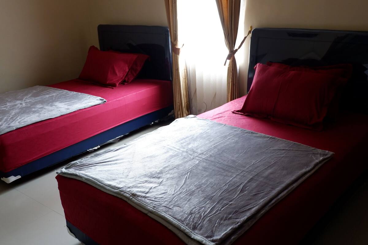 Dos camas individuales con ropa de cama roja y sábanas grises, una ventana con cortinas y una pared lisa.