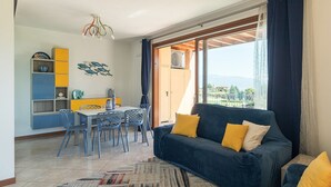 A living room with a blue sofa, yellow cushions, a dining area with a table and chairs, a bookshelf, and a view through a large window.