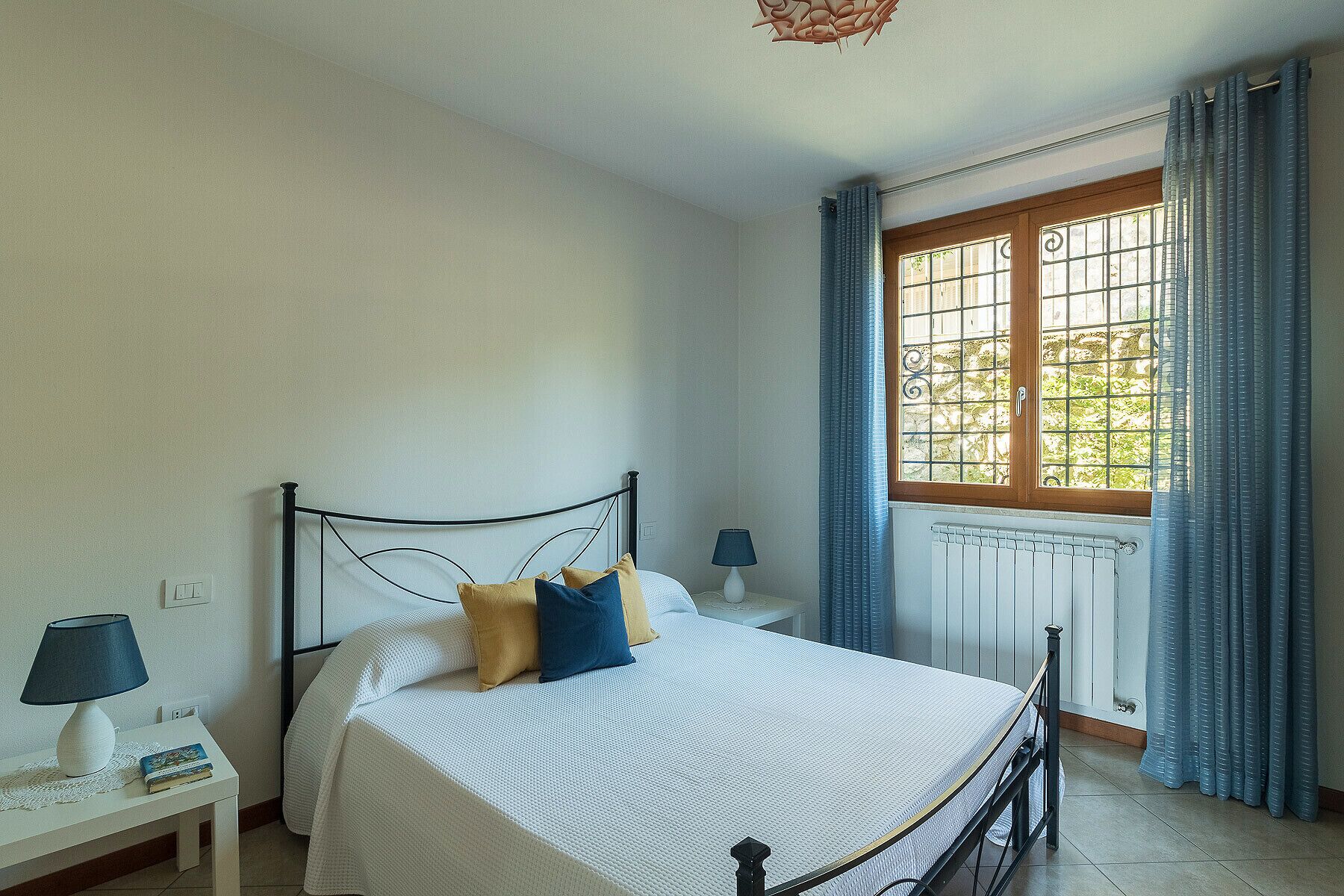 A bedroom with a bed, a nightstand, a lamp, a window with curtains, and a radiator.
