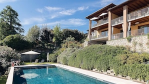 A swimming pool with lounge chairs and umbrellas, surrounded by lush greenery and a stone wall.