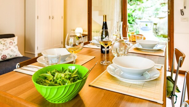 Una mesa de comedor dispuesta con platos, tazones, vasos y una botella de vino, con un tazón verde de ensalada al frente.