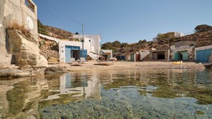 A coastal area with clear water, rocky shore, and buildings with blue doors. - The Elephant Goupa Beach (Kimolos)