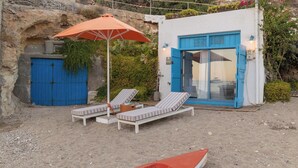 A beachside property with a blue door, a small boat, and two lounge chairs under an umbrella. - The Elephant Goupa Beach (Kimolos)