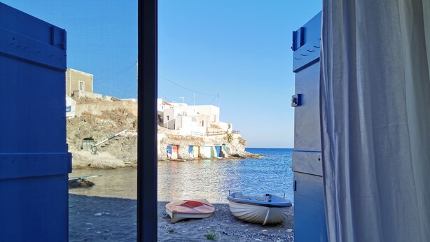 A coastal scene with a rocky shoreline, a few boats, and buildings in the background. - The Elephant Goupa Beach (Kimolos)