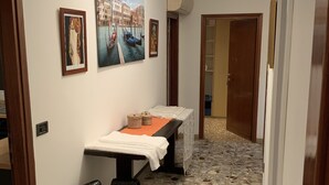 A hallway with a stone floor, a massage table, and a chair. There are framed pictures on the wall and a door at the end. - Venice Sweet Home (Venice)