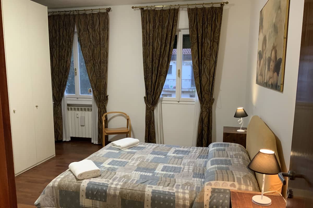 A bedroom with a bed, a chair, a nightstand with a lamp, and a window with curtains.
