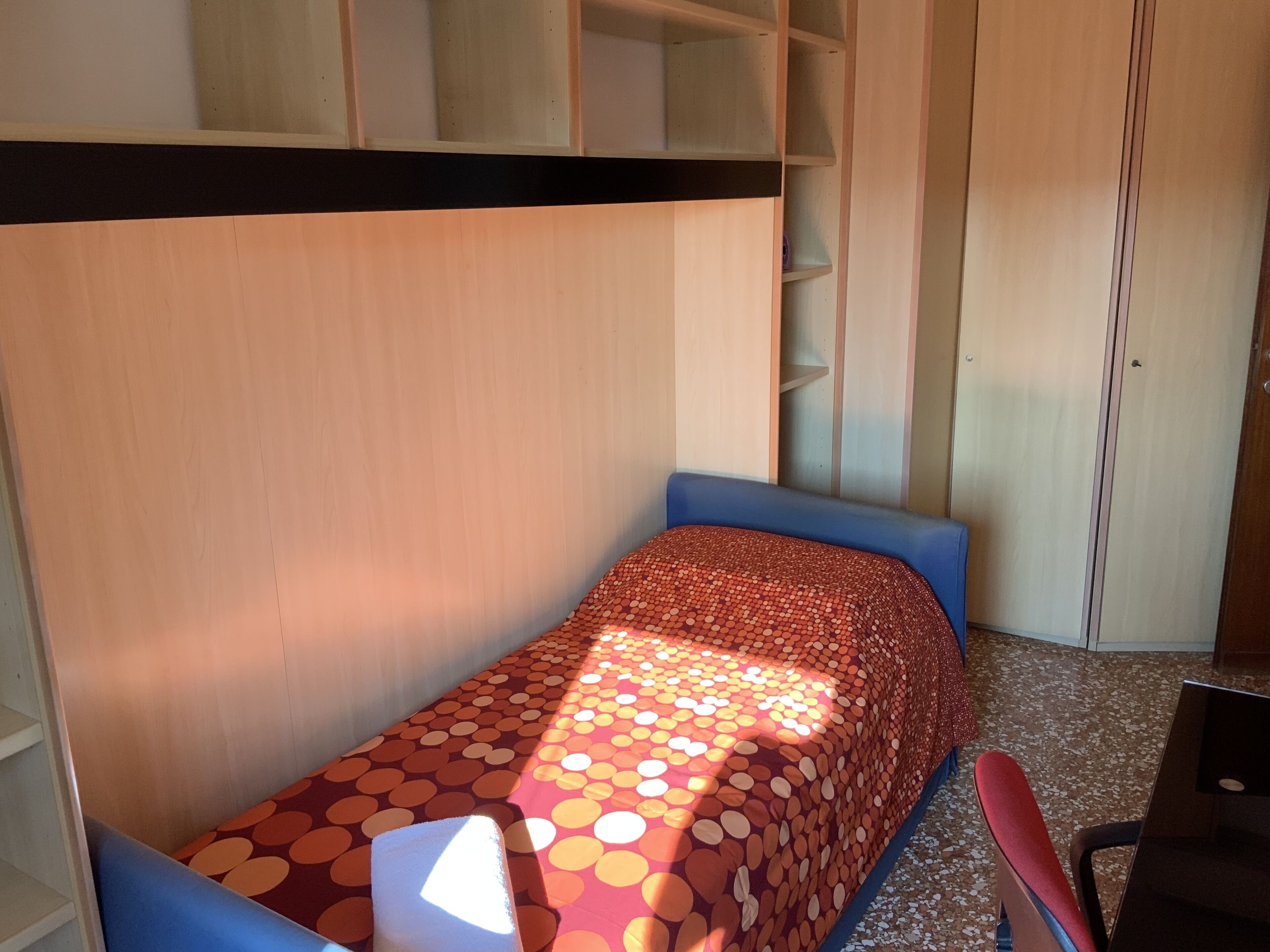 A small, single-bed room with a patterned bedspread, a desk, and a chair.