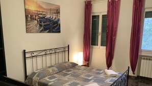 A bedroom with a bed, a nightstand, a lamp, a window with curtains, and a painting on the wall. - Venice Sweet Home (Venice)