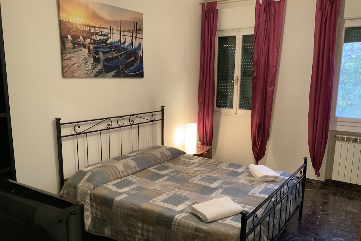 A bedroom with a bed, a nightstand, a lamp, a window with curtains, and a painting on the wall.
