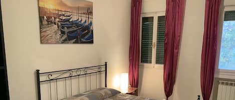 A bedroom with a bed, a nightstand, a lamp, a window with curtains, and a painting on the wall.