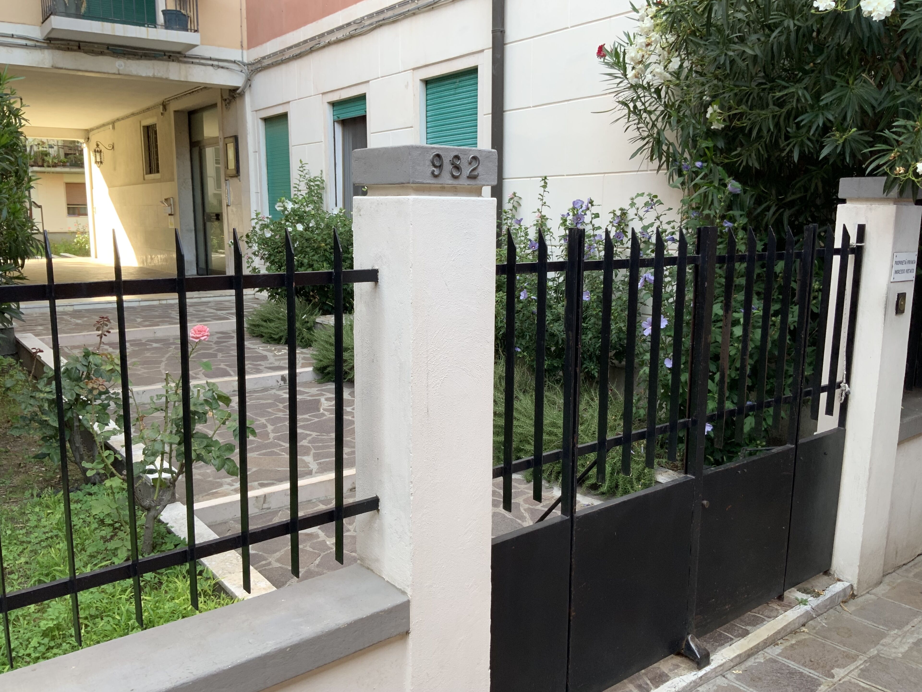 Área residencial murada com placa de número 982, caminho pavimentado e vegetação ao redor.