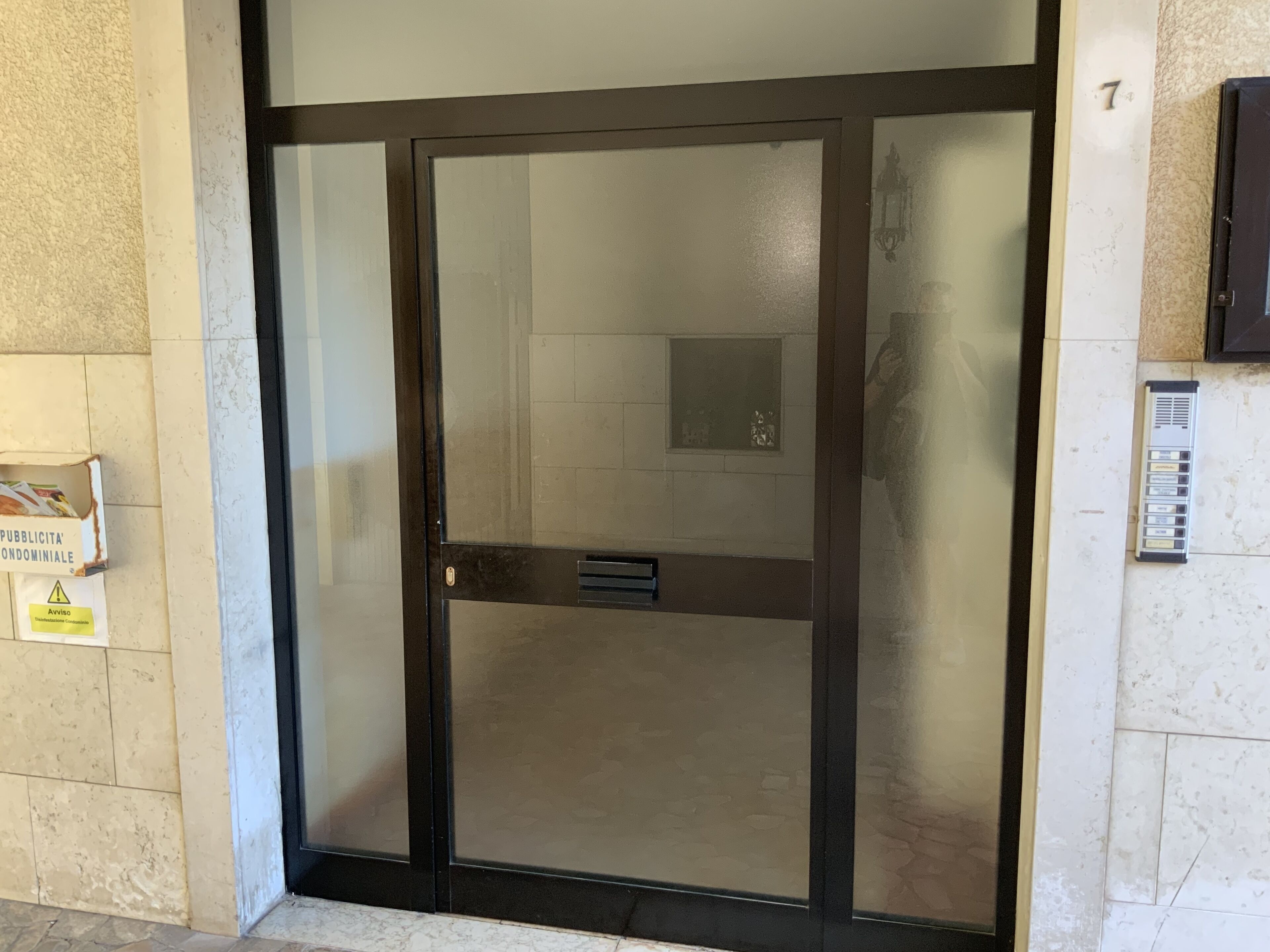 Ingresso con porta in vetro, una maniglia in metallo, una piccola bocchetta di aerazione e un sistema di intercomunicazione montato a parete.