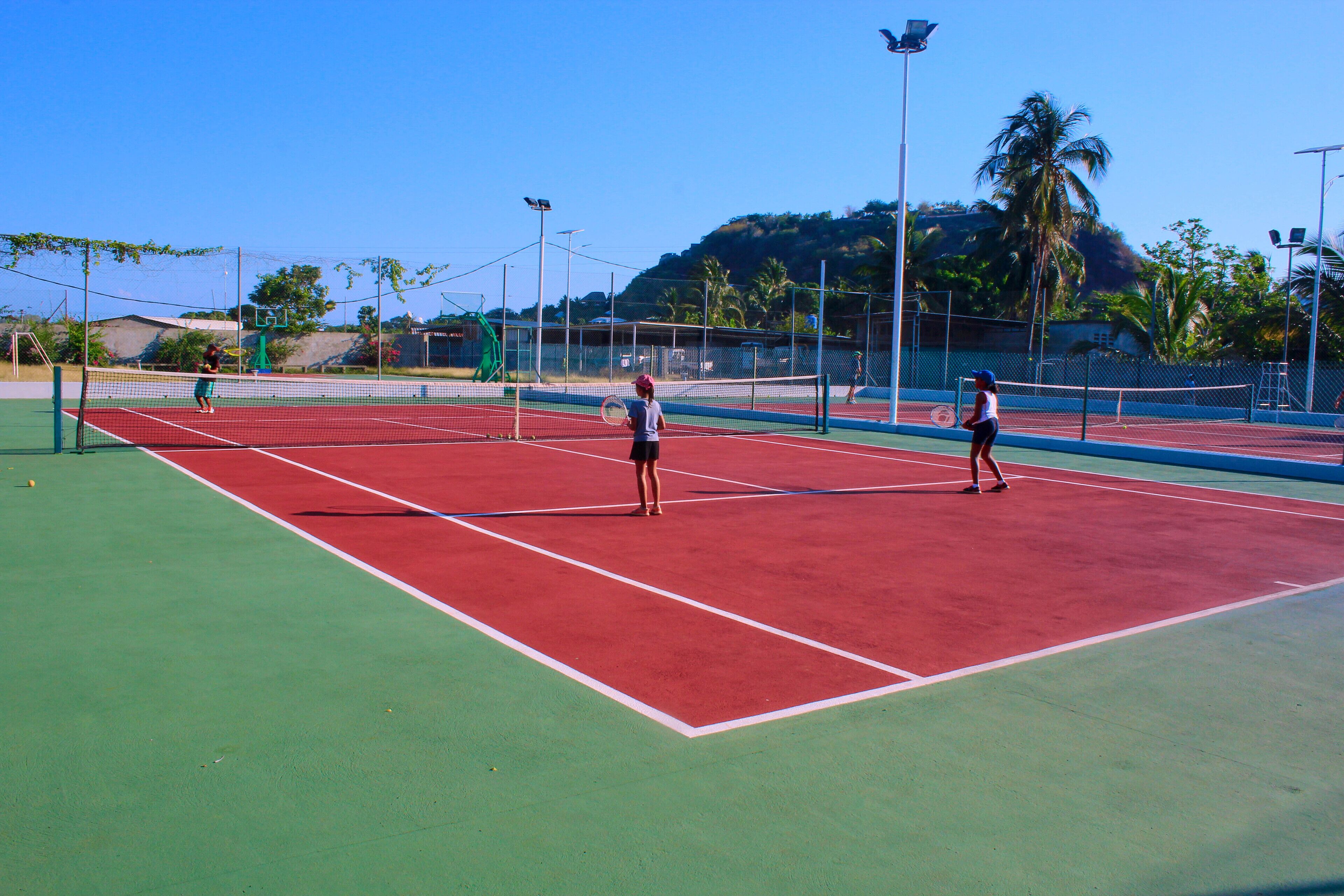 A tennis court with multiple courts, surrounded by a fence, and palm trees in the background.