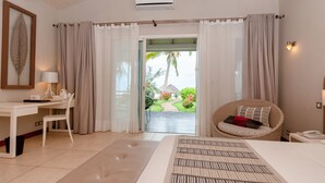 A bedroom with a bed, a desk with a chair, a wicker chair with cushions, and a view of a garden through a glass door. - Palm Beach Resort & Spa (Nosy Be)