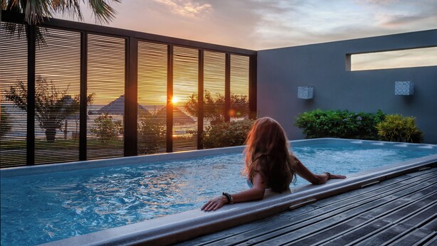 A person relaxing in a modern outdoor pool with a sunset view, surrounded by a wooden deck and a metal railing. - Palm Beach Resort & Spa (Nosy Be)