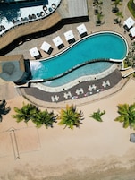 An aerial view of a resort with a large curved swimming pool, sun loungers, and palm trees.