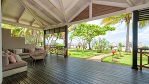 A covered outdoor seating area with a wooden deck, a sofa with cushions, and a wicker chair. - Palm Beach Resort & Spa (Nosy Be)