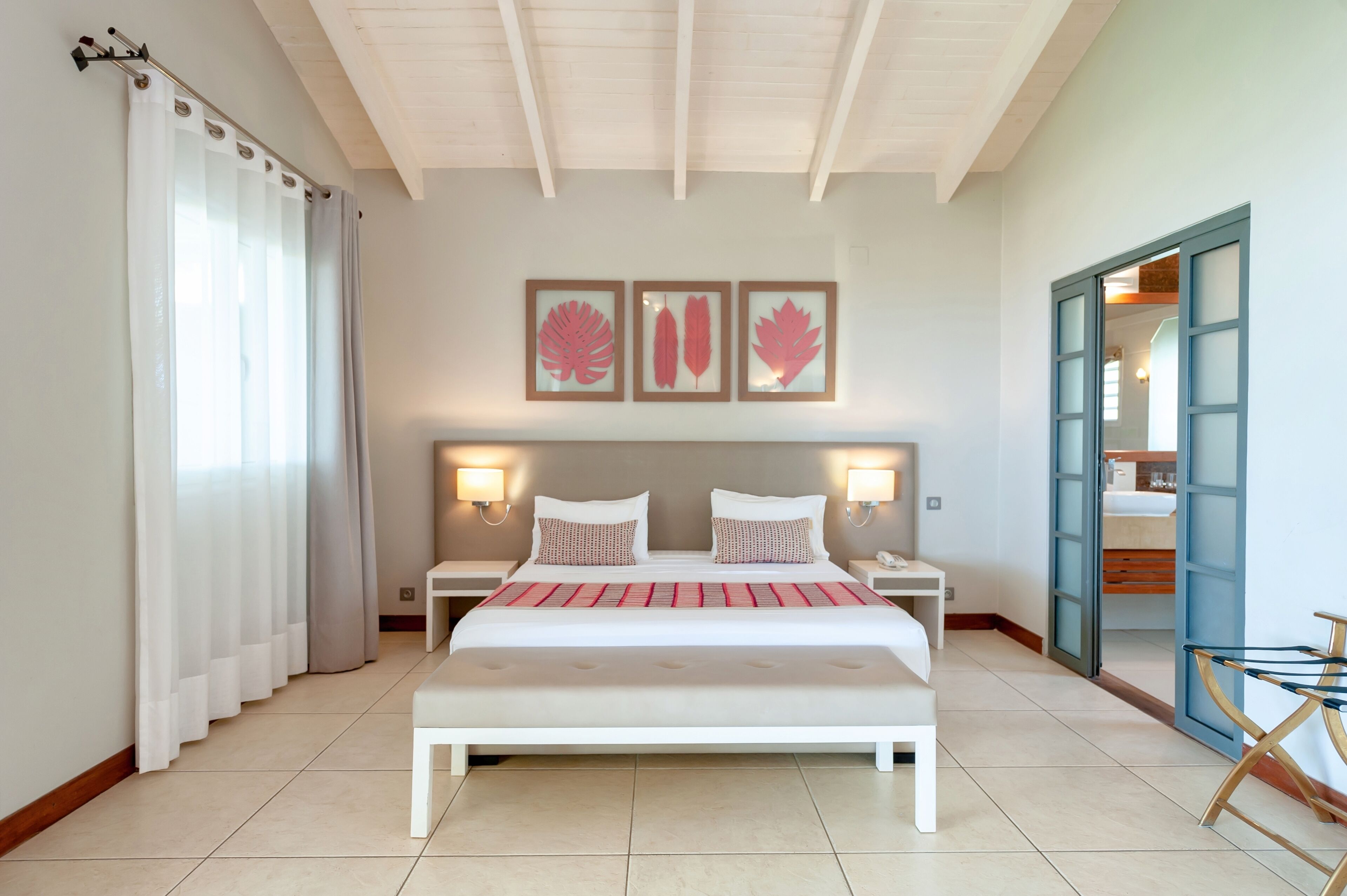 Un dormitorio con una cama grande, dos mesitas de noche, un banco, una puerta y tres cuadros en la pared.
