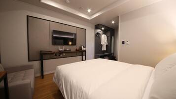 A modern hotel room with a large bed, a desk with a flat-screen TV, a robe hanging on the wall, and a small seating area.