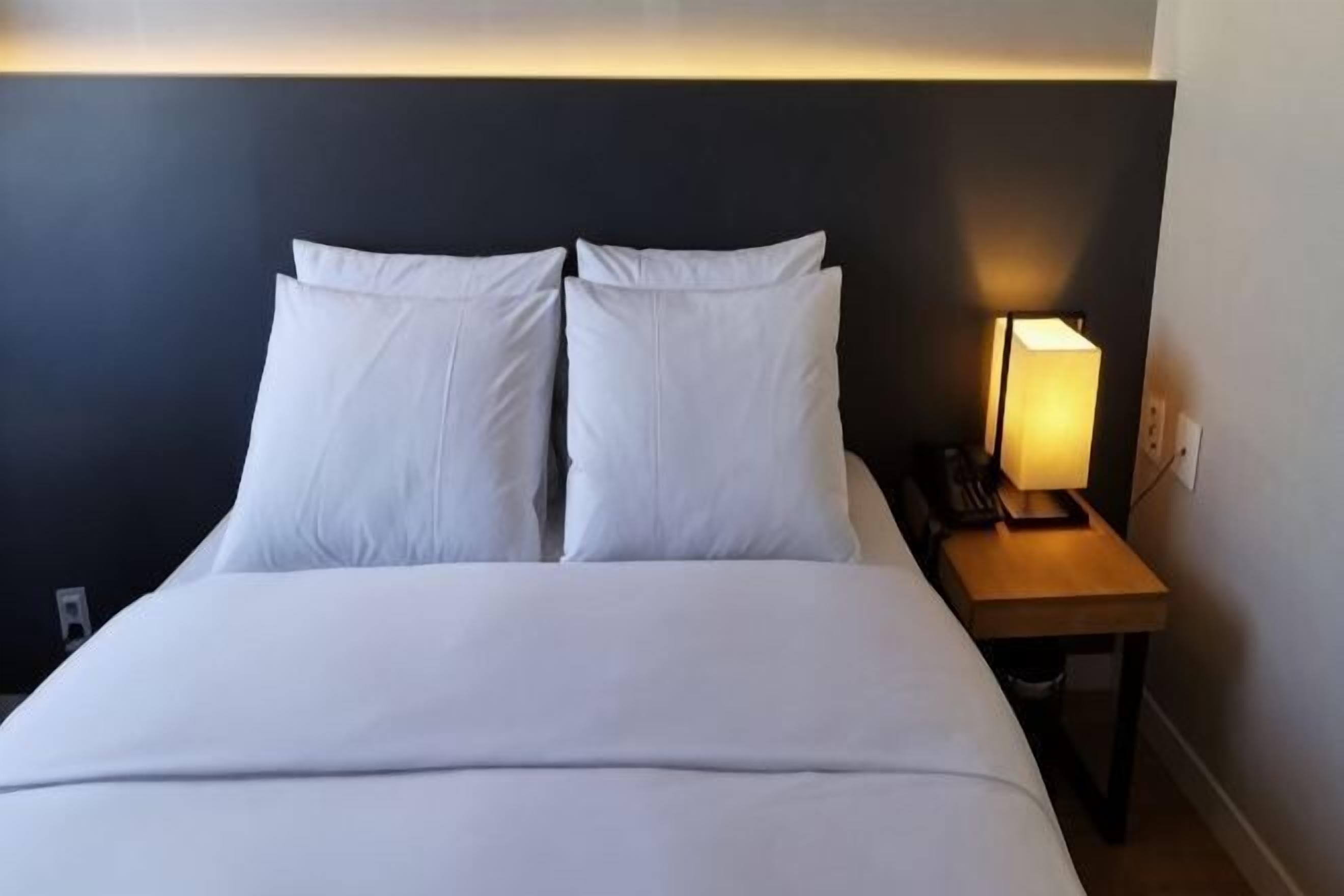 A hotel room with a bed, two pillows, a bedside table with a lamp, and a wall socket.