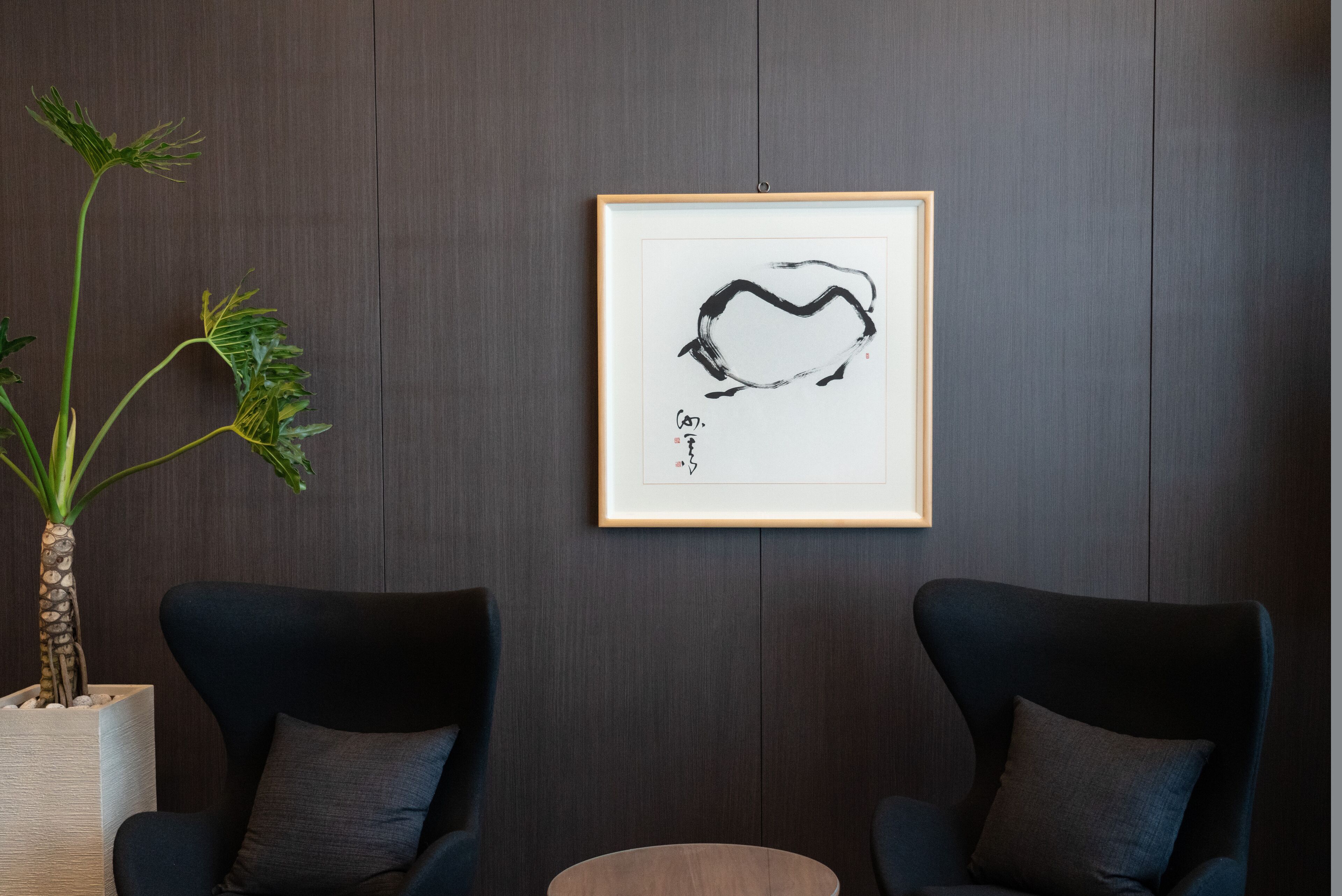 A room with two dark blue armchairs, a round wooden table, and a framed abstract artwork on the wall.
