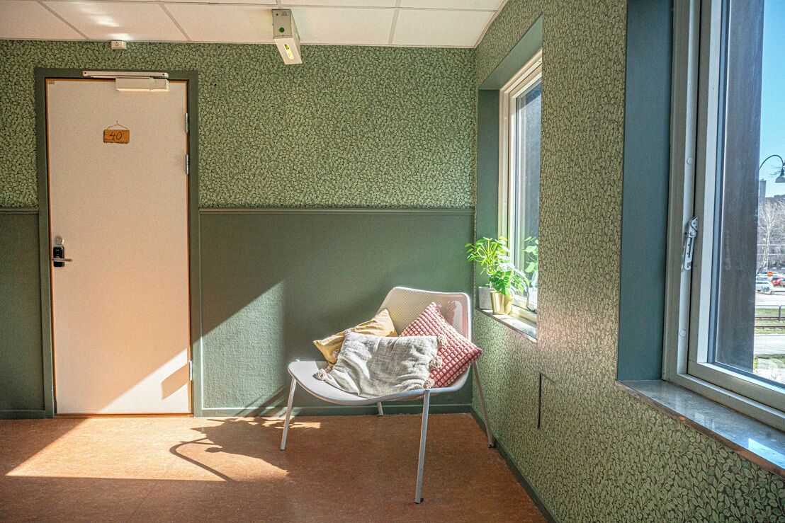 A room with a green textured wall, a white door with a number sign, a window with a view, and a chair with cushions.