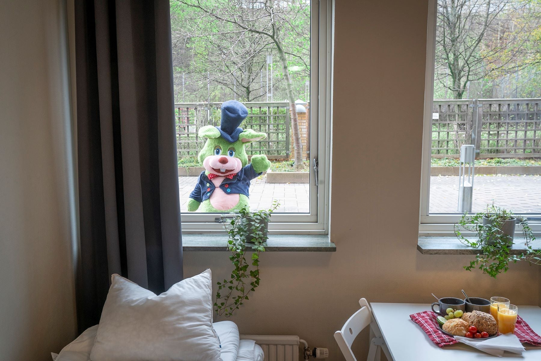 Ein Zimmer mit Fenster, Heizkörper, einem weißen Stuhl, einem Tisch mit Frühstücksgebäck und einem Stofftier vor dem Fenster.