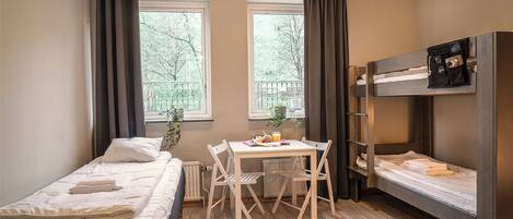 Een studentenkamer met twee eenpersoonsbedden, een stapelbed, een houten tafel met stoelen en een raam met gordijnen.