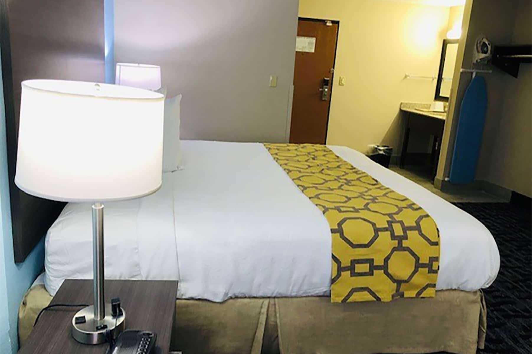 Una habitación de hotel con una cama, una lámpara de noche, una mesita con un teléfono y un baño al fondo.