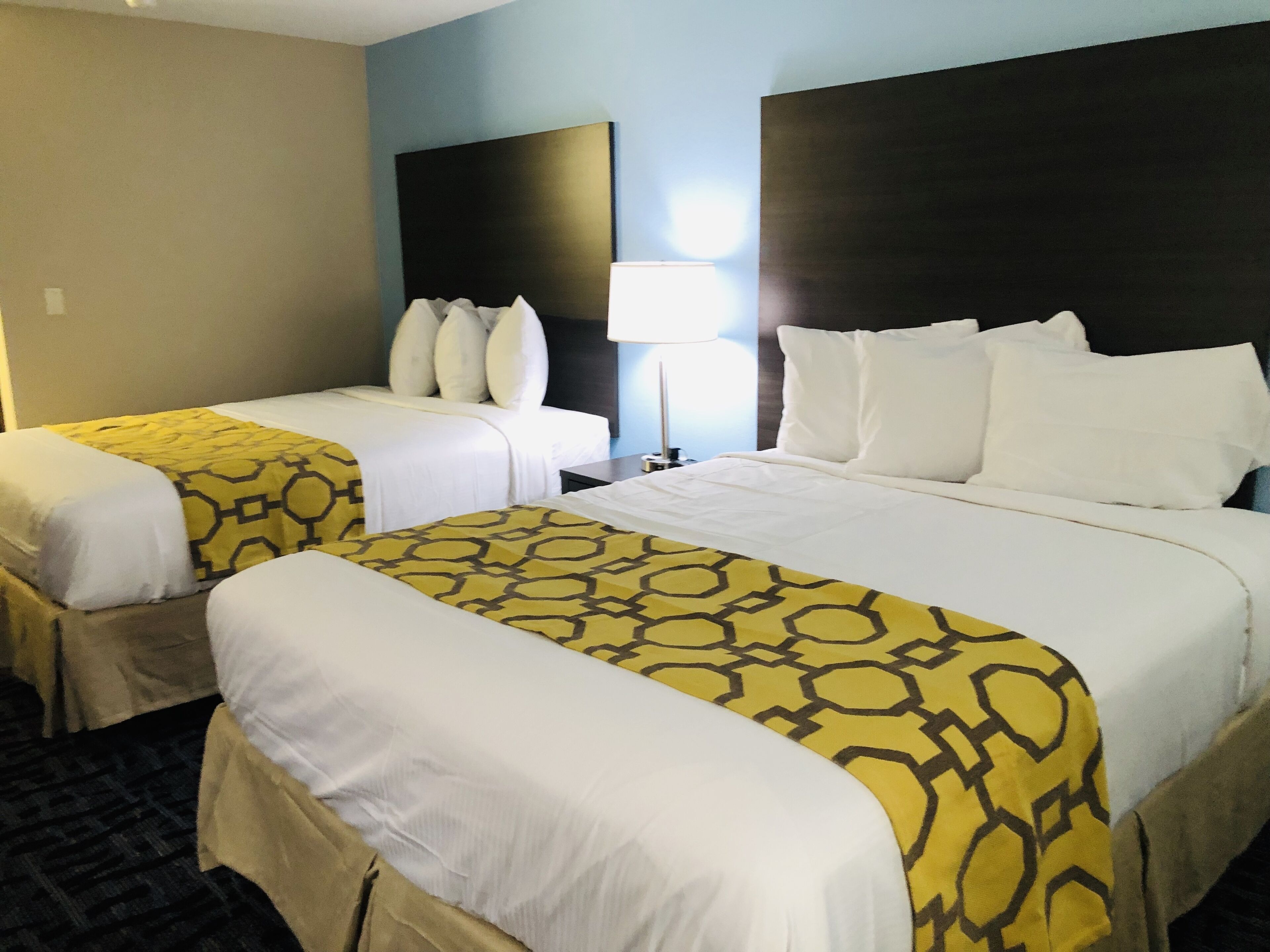 A hotel room with two beds, each with a yellow patterned bedspread and white linens. There is a nightstand with a lamp between the beds.